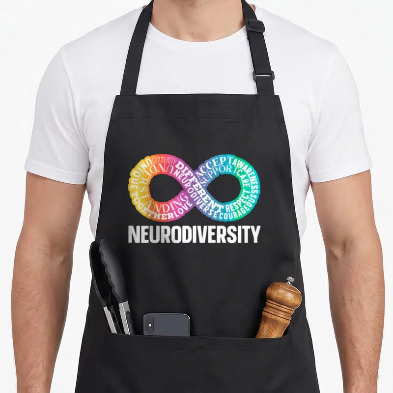 Neurodiversity neurodivergent rainbow apron - Infinity symbol formed by colorful words COMMUNITY ACCEPT AWARENESS below — shown on a black cotton canvas apron against a white background