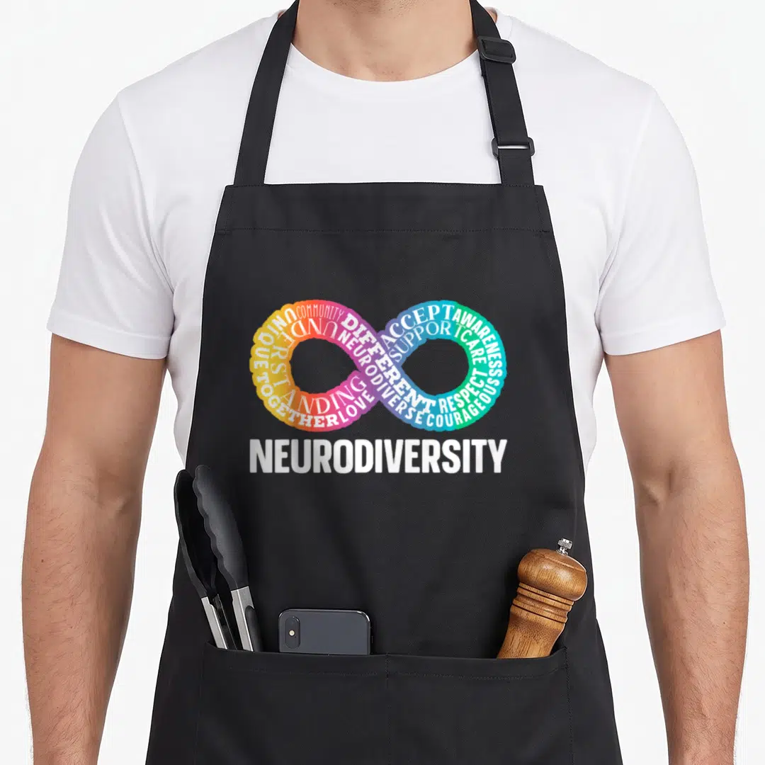 Neurodiversity neurodivergent rainbow apron - Infinity symbol formed by colorful words COMMUNITY ACCEPT AWARENESS below — shown on a black cotton canvas apron against a white background