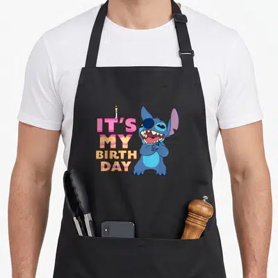 Stack stitch apron - Stacked It's My Birthday text in pink and orange with blue cartoon character and candle — shown on a black cotton canvas apron against a white background