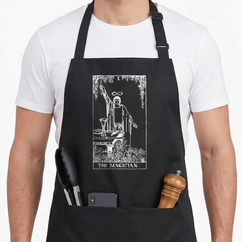 Magician traditional apron - White line art of The Magician tarot figure with floral border and text — shown on a black cotton canvas apron against a white background
