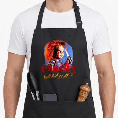Play collectible apron - Chucky doll holding knife with red CHUCKY text and yellow WANNA PLAY? script — shown on a black cotton canvas apron against a white background