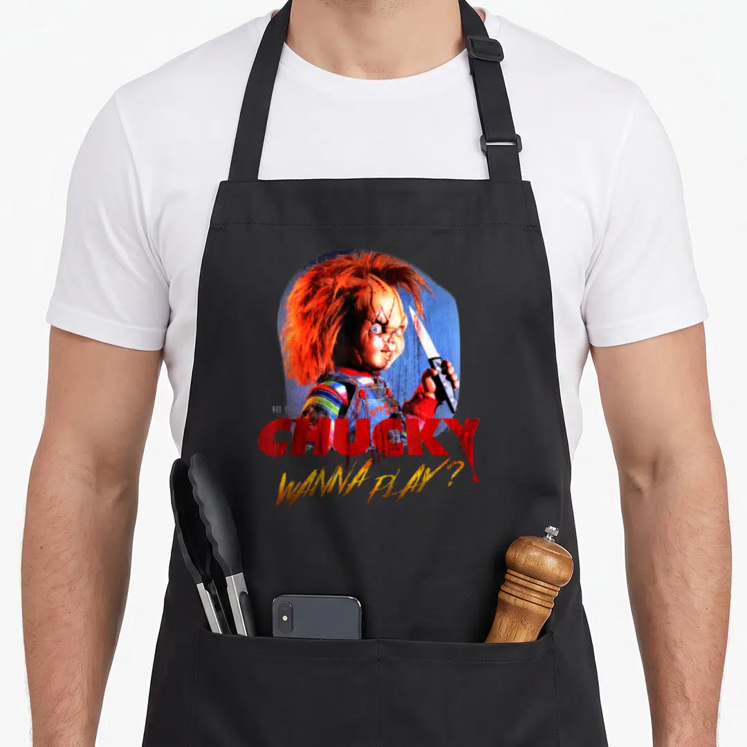 Play collectible apron - Chucky doll holding knife with red CHUCKY text and yellow WANNA PLAY? script — shown on a black cotton canvas apron against a white background