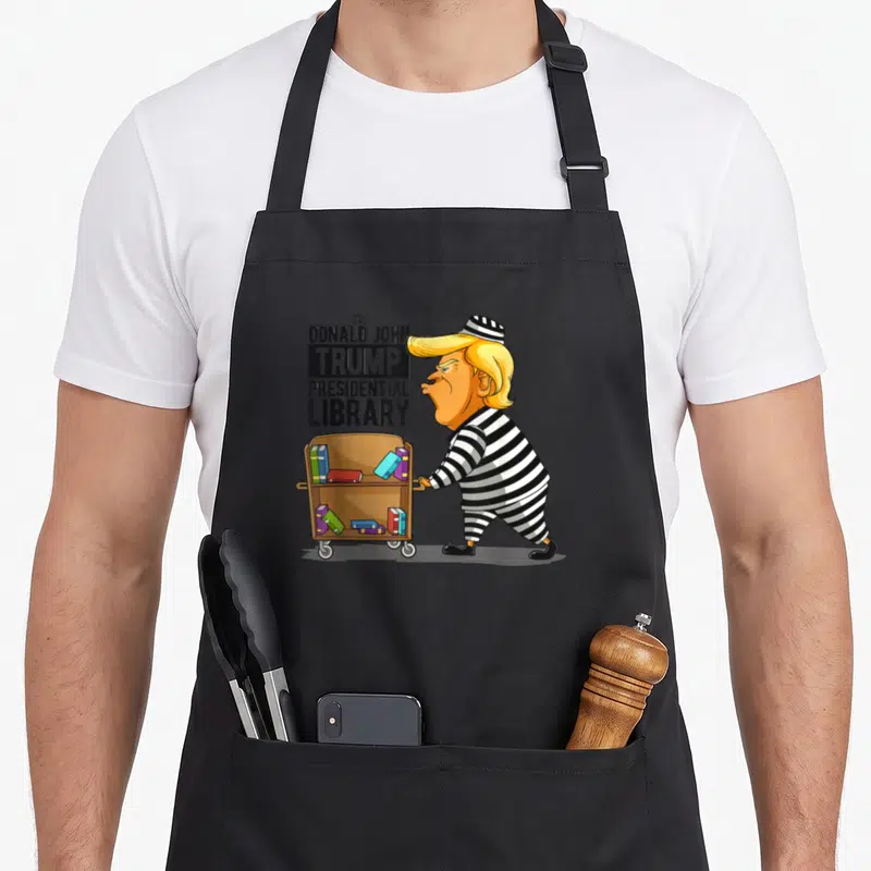 Anti crew apron - Cartoon of Trump in prison stripes pushing a book cart next to stacked text reading The Donald John — shown on a black cotton canvas apron against a white background