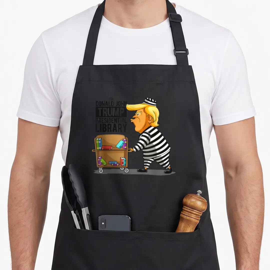 Anti crew apron - Cartoon of Trump in prison stripes pushing a book cart next to stacked text reading The Donald John — shown on a black cotton canvas apron against a white background