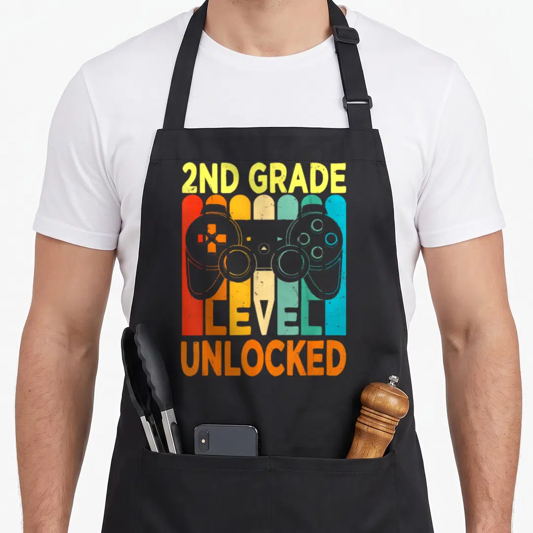 Hello video game apron - Retro controller over five colorful stripes with 2nd Grade Level Unlocked text — shown on a black cotton canvas apron against a white background