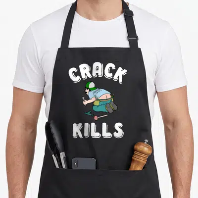 Crack plumber apron - Cartoon plumber kneeling between arched CRACK and bottom KILLS text in white, blue, green, teal — shown on a black cotton canvas apron against a white background