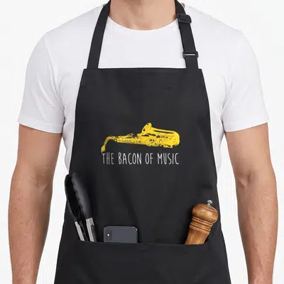Marching bacon apron - Yellow distressed saxophone above white text THE BACON OF MUSIC — shown on a black cotton canvas apron against a white background