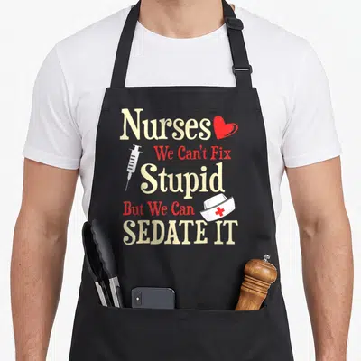 Fix nurse apron - Stacked typography reading Nurses We Can't Fix Stupid But We Can Sedate It with a heart syringe — shown on a black cotton canvas apron against a white background