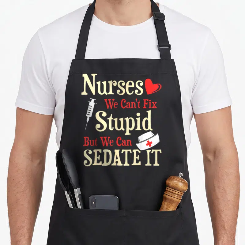 Fix nurse apron - Stacked typography reading Nurses We Can't Fix Stupid But We Can Sedate It with a heart syringe — shown on a black cotton canvas apron against a white background