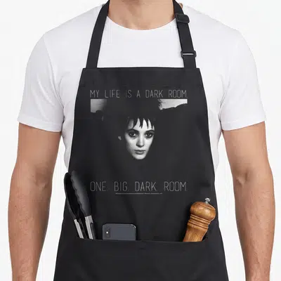 Lydia beetle apron - Woman's face with text My Life Is A Dark Room in grey and white on black background — shown on a black cotton canvas apron against a white background