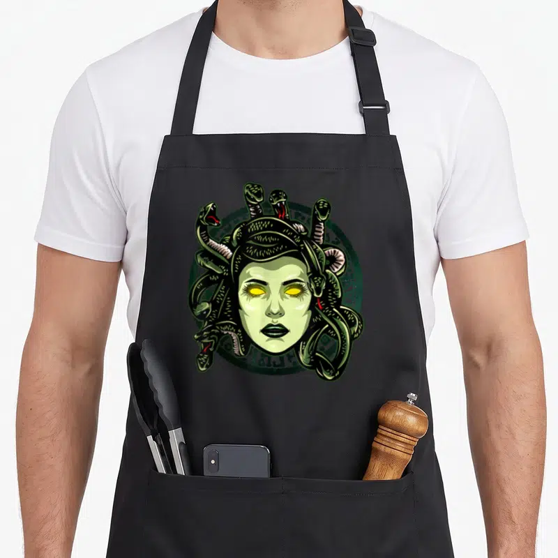 Monster greek aprons - Cartoon Medusa face with green snakes and yellow eyes on a dark green circular emblem — shown on a black cotton canvas apron against a white background