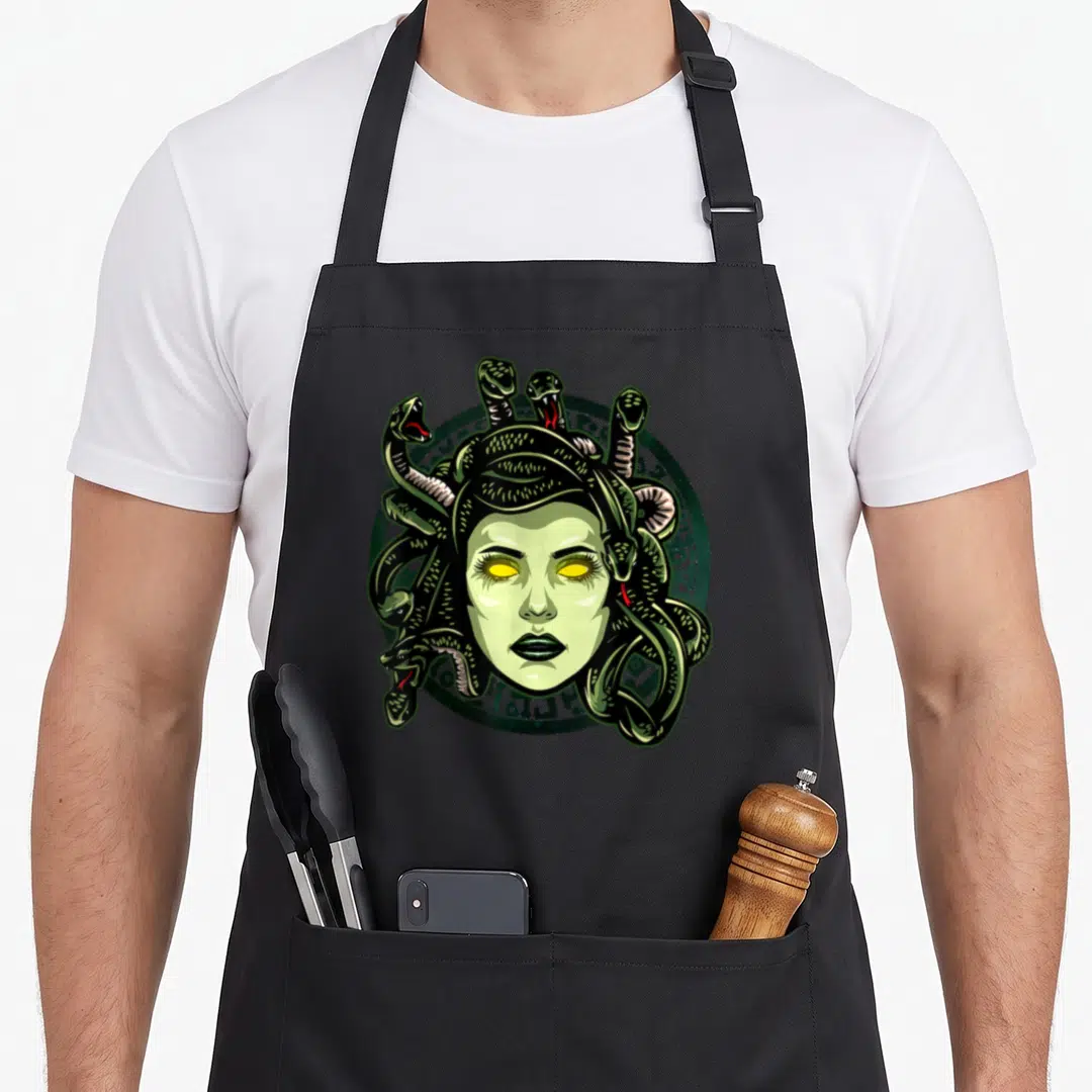Monster greek aprons - Cartoon Medusa face with green snakes and yellow eyes on a dark green circular emblem — shown on a black cotton canvas apron against a white background