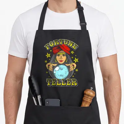 Fortune carnival apron - Cartoon fortune teller holding blue crystal ball with yellow FORTUNE TELLER text and scattered — shown on a black cotton canvas apron against a white background