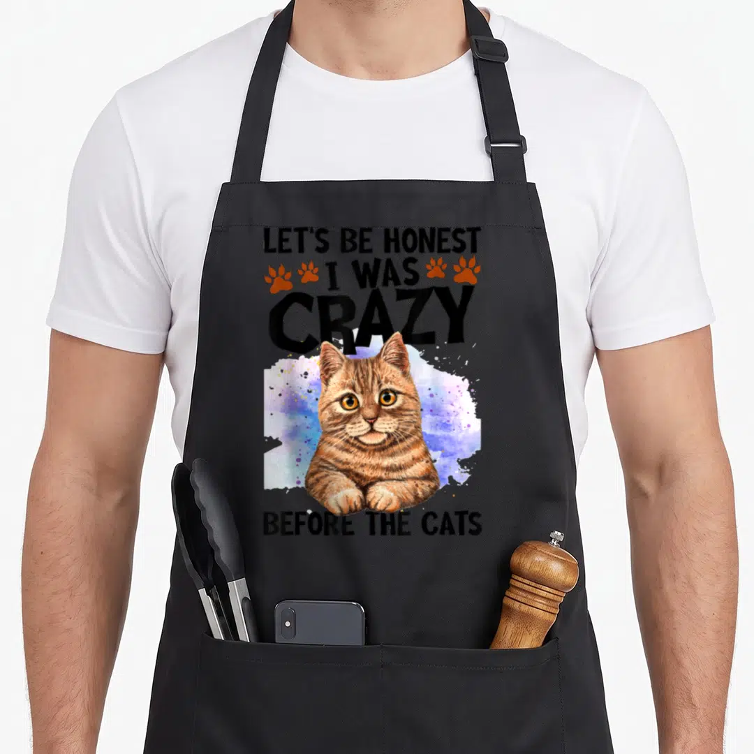 Honest black cat aprons - Cartoon brown tabby cat over blue purple orange splash with text and orange paw prints — shown on a black cotton canvas apron against a white background
