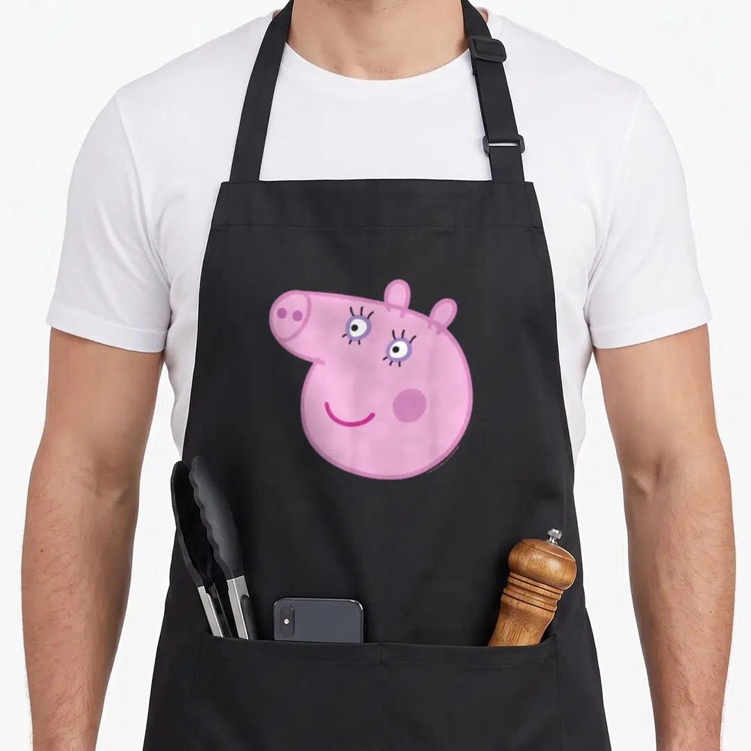 Mummy face apron - Large pink cartoon pig face with eyelashes and snout on black fabric — shown on a black cotton canvas apron against a white background