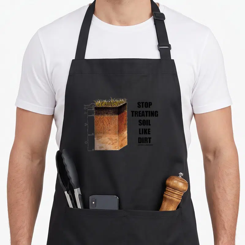 Stop science apron - Soil profile diagram with O A B C labels and stacked text STOP TREATING SOIL LIKE DIRT — shown on a black cotton canvas apron against a white background