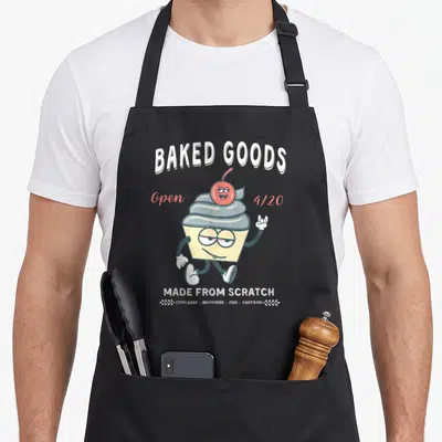 Handmade cupcake apron - Cartoon cupcake character with cherry, BAKED GOODS text, and 4/20 reference in retro colors — shown on a black cotton canvas apron against a white background