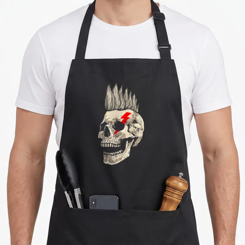 Skull punk rock apron - Beige skull with mohawk, red lightning bolt, and PUNKS NOT DEAD text — shown on a black cotton canvas apron against a white background