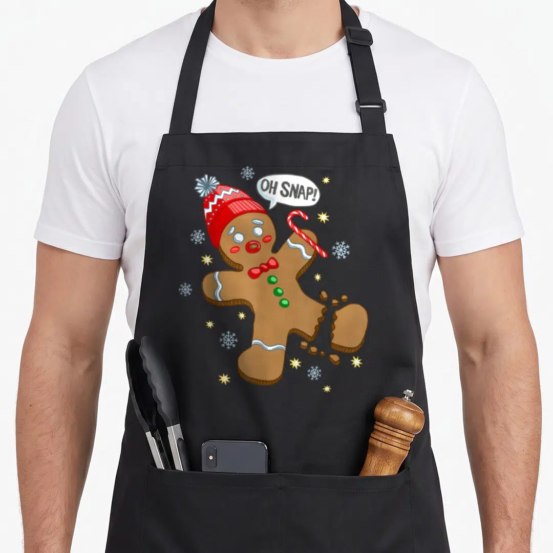 Trendy gingerbread apron - Cartoon gingerbread man with broken leg saying OH SNAP surrounded by snowflakes and stars — shown on a black cotton canvas apron against a white background