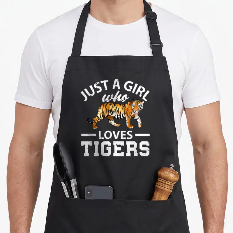 Retro tiger apron - Tiger illustration with text Just A Girl Who Loves Tigers in distressed white font — shown on a black cotton canvas apron against a white background