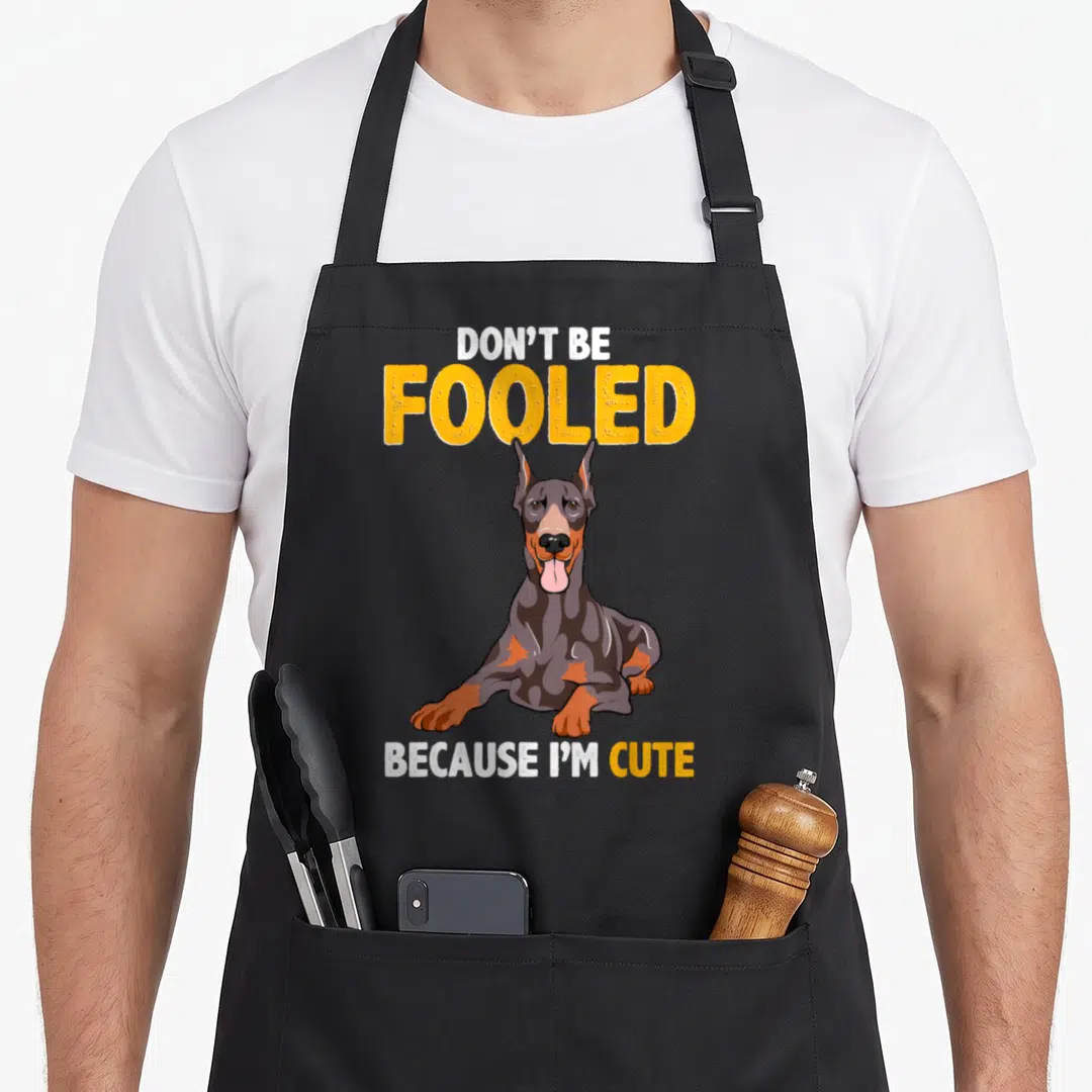 Fooled meme apron - Cartoon grey and tan Doberman sitting under DON'T BE FOOLED BECAUSE I'M CUTE text — shown on a black cotton canvas apron against a white background