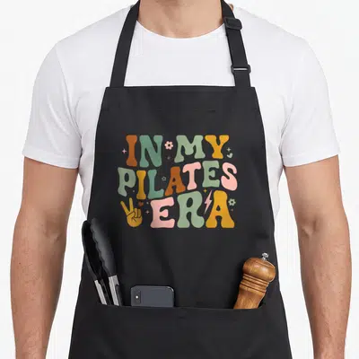 Pilate grill instructor apron - Retro bubbly text reading IN MY PILATES ERA in orange, green, yellow, and pink — shown on a black cotton canvas apron against a white background