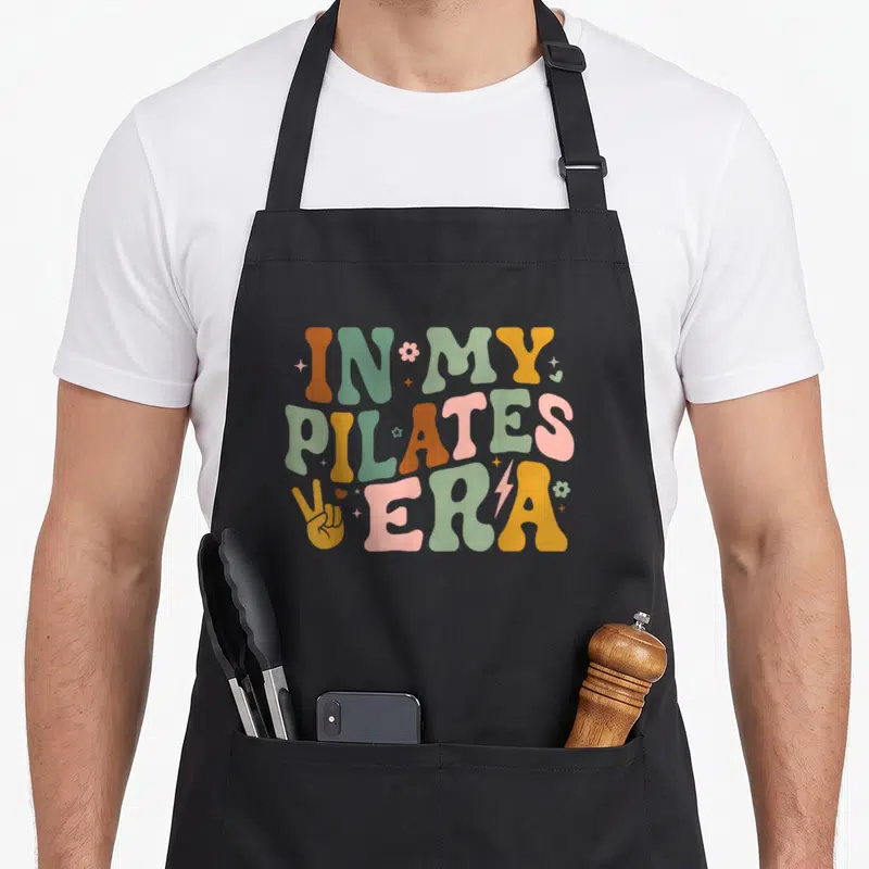Pilate grill instructor apron - Retro bubbly text reading IN MY PILATES ERA in orange, green, yellow, and pink — shown on a black cotton canvas apron against a white background