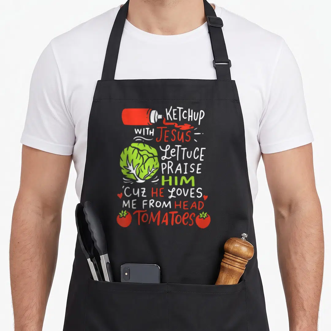 Ketchup veggie apron - Vertical text KETCHUP WITH JESUS with ketchup bottle lettuce and tomato illustrations in red — shown on a black cotton canvas apron against a white background