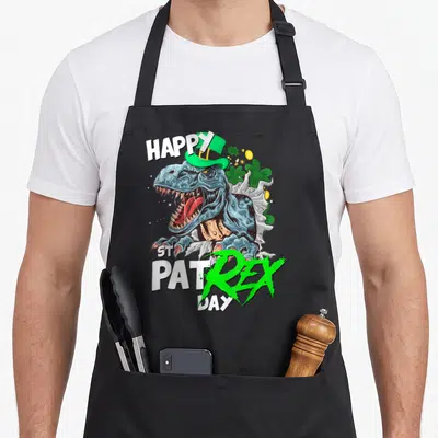 Rex st patrick day aprons - T-Rex in green hat with shamrocks and HAPPY ST PAT REX DAY text — shown on a black cotton canvas apron against a white background