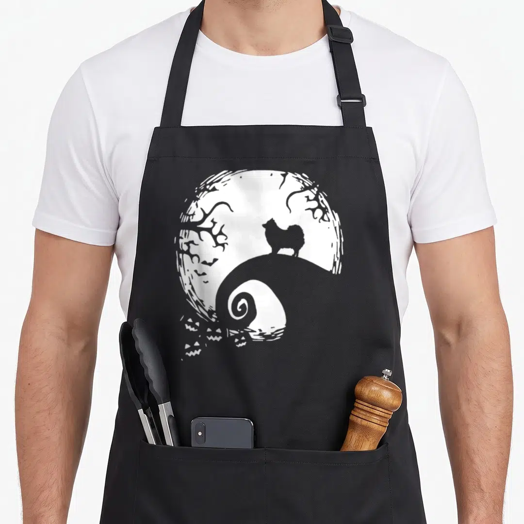 Pomeranian halloween apron - White line-art dog silhouette on spiral hill in front of large moon with pumpkins — shown on a black cotton canvas apron against a white background