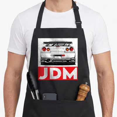 Turbo design imports vintage apron - Distressed rear car drawing above red JDM text block in white red black — shown on a black cotton canvas apron against a white background