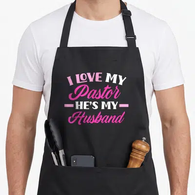 Pastor husband wife aprons - Stacked pink and white text reading I Love My Pastor He's My Husband with horizontal lines — shown on a black cotton canvas apron against a white background