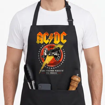 Those ac dc apron - AC/DC logo, 1981 cannon in red circle, yellow lightning bolt, white stars, and stacked text — shown on a black cotton canvas apron against a white background