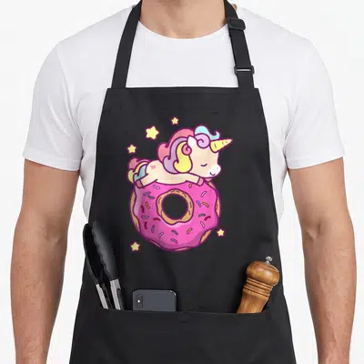 Unicorn donut apron - Lying cartoon unicorn on pink frosted donut with five yellow stars and rainbow mane — shown on a black cotton canvas apron against a white background