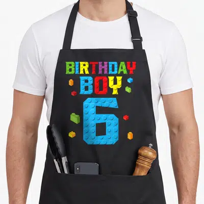 6th grill master apron - BIRTHDAY BOY 6 text in colorful brick style with scattered small plastic bricks — shown on a black cotton canvas apron against a white background