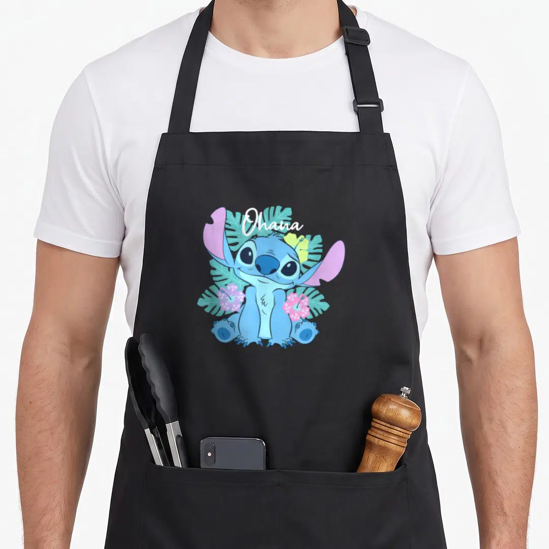 Ohana floral apron - Blue Stitch sitting amid teal leaves and pink flowers under white Ohana script — shown on a black cotton canvas apron against a white background