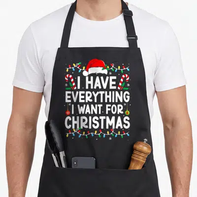 Christmas apron for dad - I Have Everything I Want For Christmas text with Santa hat candy canes and lights — shown on a black cotton canvas apron against a white background
