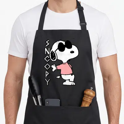 Funny joe cool apron - Snoopy in pink shirt and sunglasses next to vertical SNCOPY text in white black and pink — shown on a black cotton canvas apron against a white background