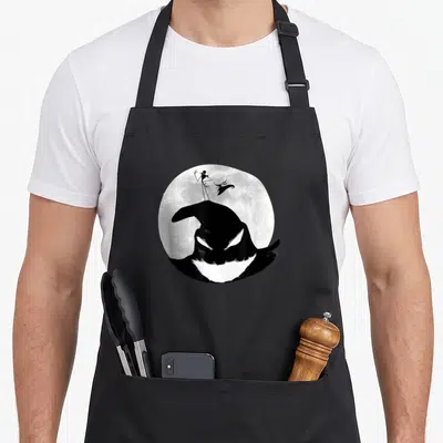 Oogie christmas apron - Oogie Boogie head silhouette under a moon with two small figures in black and white — shown on a black cotton canvas apron against a white background