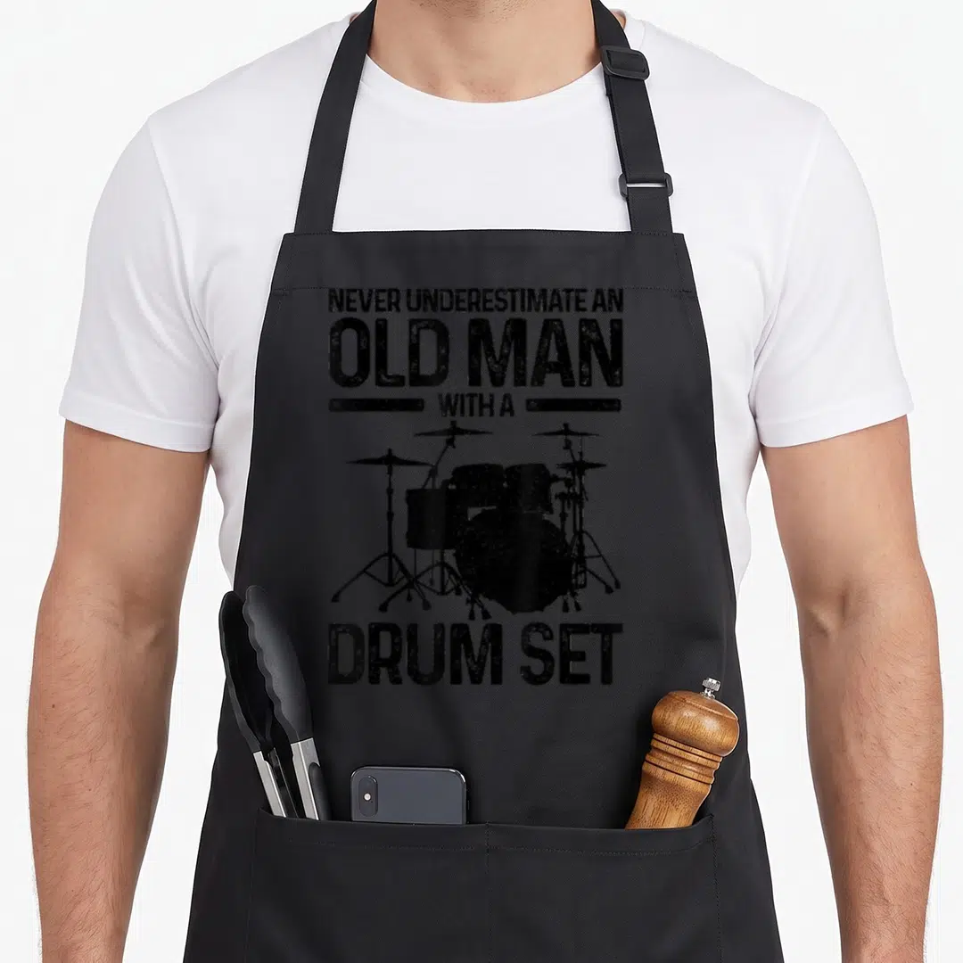 Drummer drum corps apron - Distressed black text reading NEVER UNDERESTIMATE AN OLD MAN WITH A DRUM SET above a drum — shown on a black cotton canvas apron against a white background