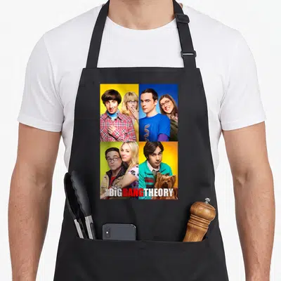 Vintage shot box apron - Cast photo split into yellow blue green orange quadrants with Big Bang Theory text and atom — shown on a black cotton canvas apron against a white background