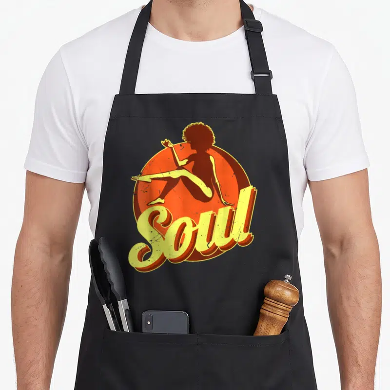 Music 70s apron - Retro woman silhouette sitting before orange circle with yellow Soul script text — shown on a black cotton canvas apron against a white background