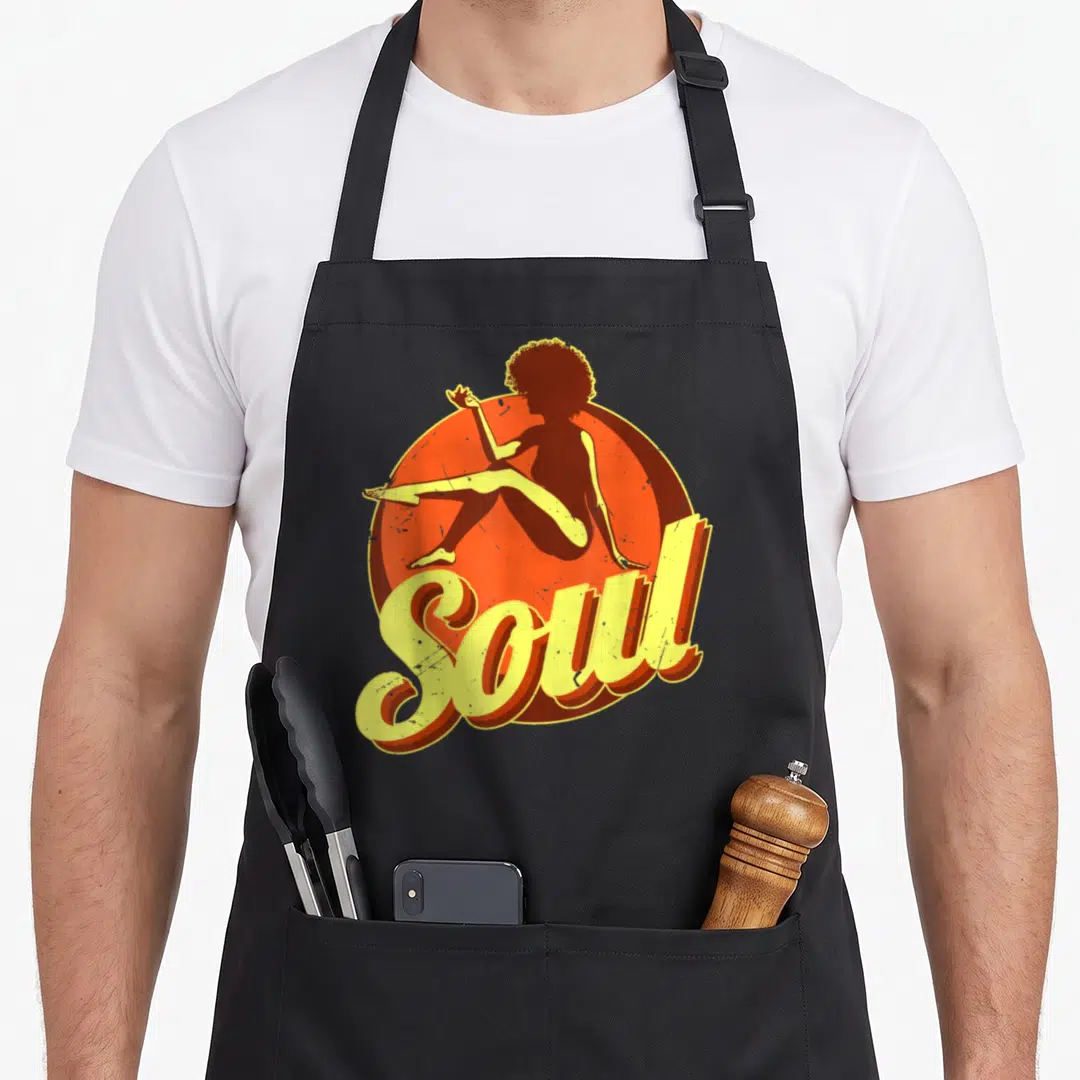 Music 70s apron - Retro woman silhouette sitting before orange circle with yellow Soul script text — shown on a black cotton canvas apron against a white background