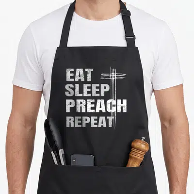 Eat youth pastor apron - Distressed white text reading EAT SLEEP PREACH REPEAT with a cross graphic on a black — shown on a black cotton canvas apron against a white background