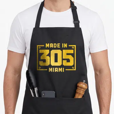 Area pride apron - Yellow text reading Made In 305 Miami inside a rectangular frame with corner screws — shown on a black cotton canvas apron against a white background