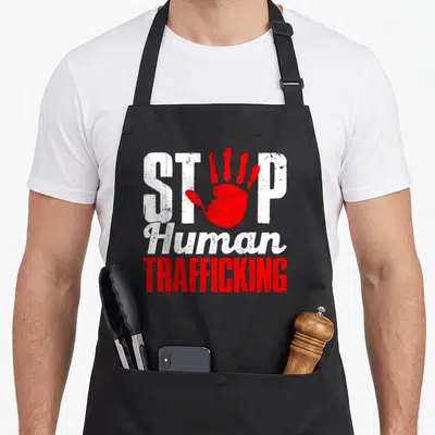 Human modern apron - Red handprint overlapping white STOP text above Human TRAFFICKING in distressed typography — shown on a black cotton canvas apron against a white background