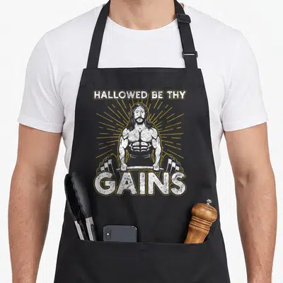Hallowed gains apron - Distressed white muscular man holding barbell with gold sunburst and text HALLOWED BE THY GAINS — shown on a black cotton canvas apron against a white background