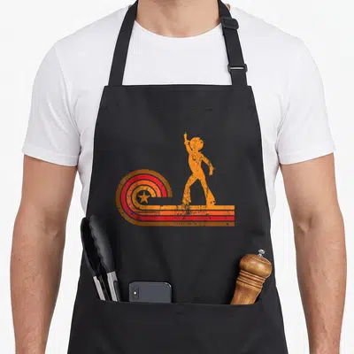 Disco 1970s apron - Orange dancing silhouette beside a distressed red yellow and orange rainbow arc with a central star — shown on a black cotton canvas apron against a white background