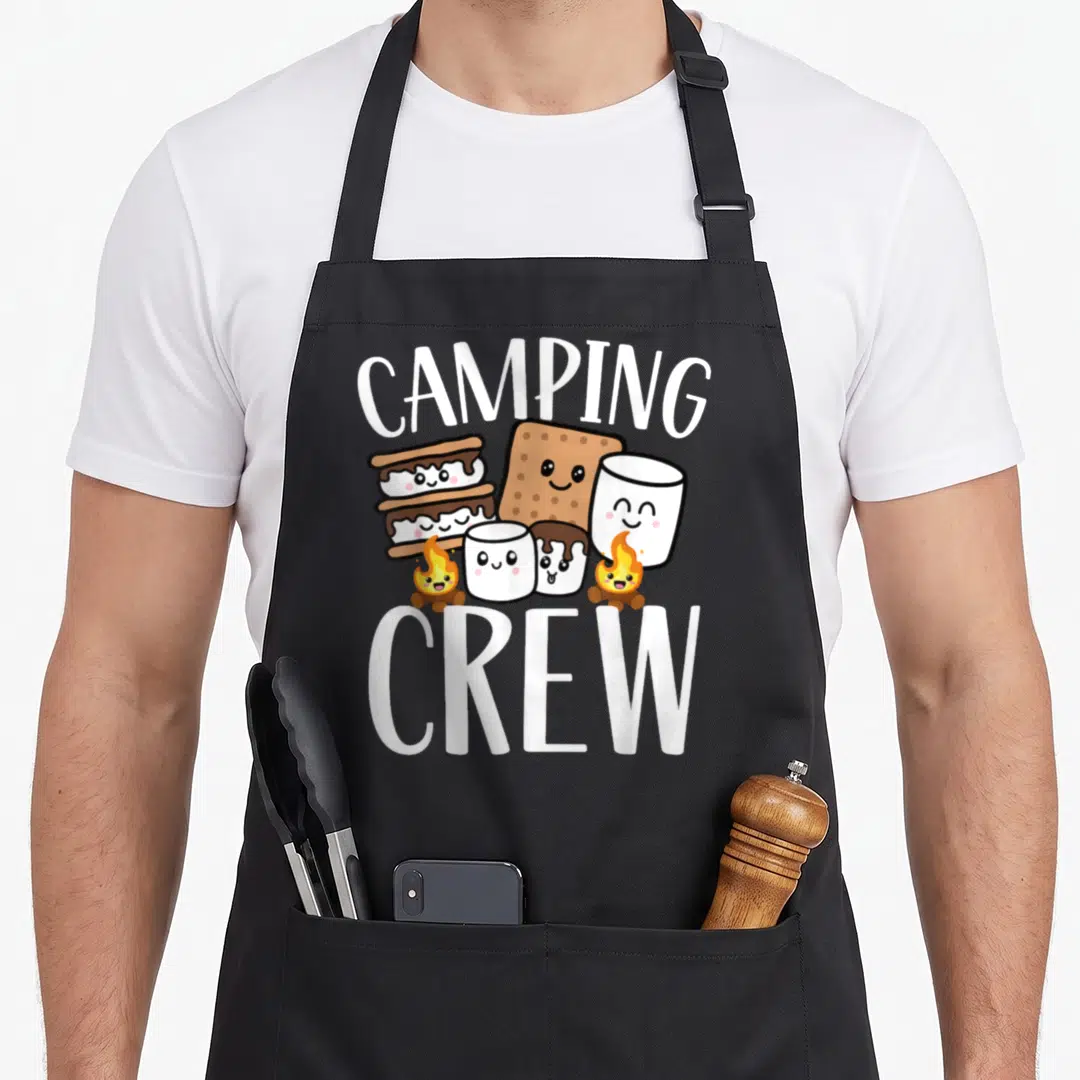 Crew smore camping apron - Arched CAMPING CREW text around smiling s'mores characters and campfires in white brown — shown on a black cotton canvas apron against a white background