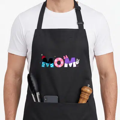Cute birthday mother's day aprons - MOM text with red hat, floral O, pink bow, peace sign hand in cartoon style using — shown on a black cotton canvas apron against a white background